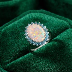 May include: An oval-shaped opal ring featuring a central iridescent stone with flashes of pink, orange, and green. The opal is surrounded by a halo of small, round, light blue gemstones set in a metallic band, creating a vibrant contrast.