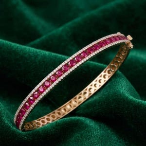 Puede incluir: Una pulsera rígida de oro con bisagra que presenta una fila central de rubíes redondos de color rojo intenso, flanqueada por dos filas delgadas de pequeños diamantes blancos transparentes. La banda metálica muestra un patrón calado.