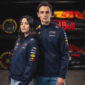 Puede incluir: Un hombre y una mujer con chaquetas azul marino con cremallera y logotipos de patrocinadores, incluyendo "Oracle Racing Honda" y "Red Bull". Llevan vaqueros azules. El fondo presenta la marca Red Bull y coches de carreras.