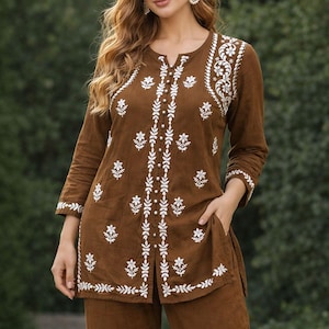 May include: A brown two-piece outfit with detailed white floral embroidery. The long-sleeved top has a split neckline and side pockets, paired with matching trousers. The ensemble is made of a soft, flowing fabric, ideal for a relaxed yet stylish look.