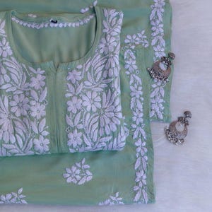 Può includere: Un completo tunica e pantaloni verde chiaro ricamato con intricati motivi floreali bianchi. Due orecchini argentati ornati con piccoli dettagli di perle sono posizionati vicino. Il tessuto appare morbido e leggero, adatto ai climi caldi.