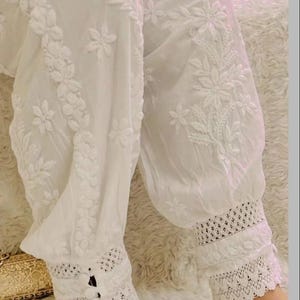 Puede incluir: Pantalones blancos bordados con motivos florales y de hojas. Los pantalones presentan un ribete de encaje en los tobillos y están hechos de una tela ligera. El diseño incluye detalles de botones en los puños de los tobillos.