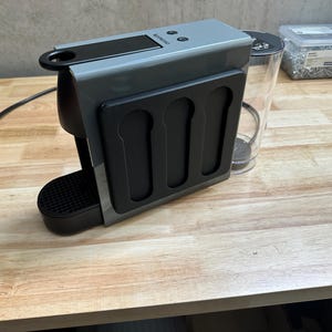 Peut inclure: Une machine à café Nespresso grise dotée d'un panneau latéral noir avec trois fentes verticales pour capsules, un bac d'égouttage noir, un réservoir d'eau transparent à l'arrière et un cordon d'alimentation noir avec une prise sur le côté.