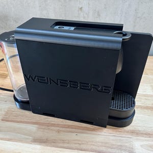 Puede incluir: Una cafetera negra con el texto WEINSBERG en relieve en el panel frontal. El aparato incluye un depósito de agua cilíndrico transparente en el lado izquierdo, una bandeja de goteo negra con patrón de rejilla y un cable de alimentación.