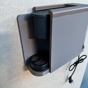 Puede incluir: Un soporte de pared para cafetera con acabado gris mate, diseñado para sostener de forma segura una cafetera de cápsulas negra y delgada. La unidad cuenta con un panel trasero plano para fijación a la pared y una base robusta con un hueco circular.