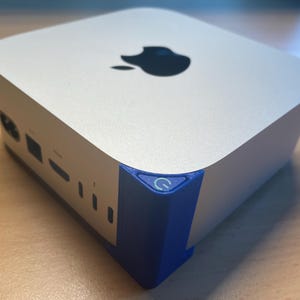 Puede incluir: Un ordenador plateado y blanco con un logotipo de Apple negro en la parte superior. La esquina presenta un acento azul con un icono de botón de encendido. Varios puertos son visibles en el lateral.