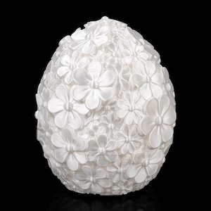May include: A white, egg-shaped decorative object covered in small, three-dimensional flowers. The flowers are tightly packed together, creating a textured surface. The background is black, highlighting the object's shape and floral design.