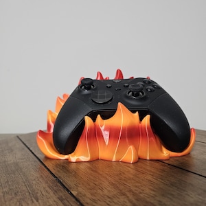May include: A black video game controller rests on a 3D-printed stand shaped like flames. The stand transitions from orange to red, creating a fiery effect. The controller has a sleek, modern design.