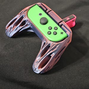 May include: A custom gaming controller grip in purple, red, and green. The grip holds a green controller with black buttons. A pink accessory is attached to the side.