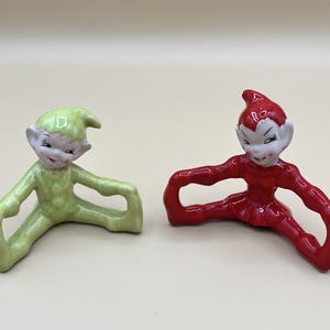 May include: Two ceramic elf figurines in a seated position with legs extended outward. One elf is light green and the other is bright red. Both figures have painted facial features, pointed hats, and arms reaching out to the sides.