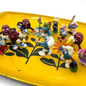 May include: A collection of small plastic figurines including blue Smurf characters and brown raisin-like characters arranged on a yellow metal tray. The tray features a black and orange floral pattern. The figures are depicted in various poses.