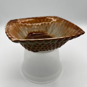 May include: A ceramic bowl with a square shape and a textured, scalloped rim. The interior has a brown and tan glaze, and the exterior has a green and brown textured pattern. The bowl is elevated on a white plastic cup.