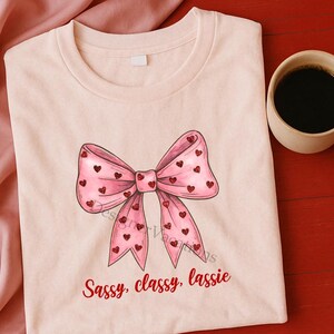 Könnte beinhalten: Hellrosa T-Shirt mit einer großen rosa Schleife mit roten Herzakzenten. Der Text "Sassy, classy, lassie" ist in roter Schrift unter der Schleife aufgedruckt. Rechts vom T-Shirt ist eine Kaffeetasse zu sehen.
