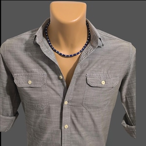 Puede incluir: Un collar de cuentas azul oscuro y plateado en un maniquí con una camisa gris abotonada. El collar está compuesto por pequeñas cuentas azul oscuro y plateadas. La camisa tiene dos bolsillos delanteros y botones blancos.