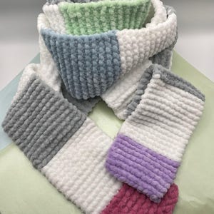 Puede incluir: Una bufanda suave y colorida con una apariencia texturizada y afelpada. La bufanda presenta secciones grises, blancas, azul claro, verdes, moradas y rosas. La bufanda está doblada y dispuesta para mostrar los bloques de color.