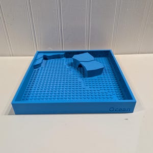 May include: A blue plastic toy with a square base and a textured surface. The toy has raised areas resembling landforms and the word "Ocean" printed on the side. The base is covered in small, circular studs.