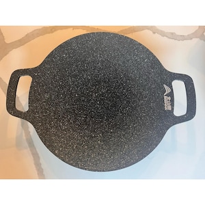 May include: A round, black speckled cooking griddle with two rectangular handles. The griddle has a textured surface and a logo on the right side. The griddle is designed for cooking.