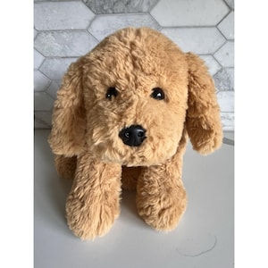 May include: A plush, golden-brown dog toy with floppy ears and a black nose. The stuffed animal has black eyes and a soft, textured fur. The toy is sitting upright, with its front paws visible. The background is a white and gray hexagon tile pattern.