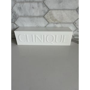 May include: A rectangular white plastic storage container featuring the brand name CLINIQUE embossed in large capital letters across the front face. The container has a smooth, matte finish and a clean, minimalist design with sharp, defined edges.