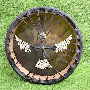 Könnte beinhalten: Eine runde, handgefertigte Trommel mit einem dunkelbraunen Lederrahmen und einem naturfarbenen Trommelfell. Schwarz-weiße Webakzente bilden ein kreuzartiges Design in der Mitte. Der Text "UBIcrafts" ist sichtbar.