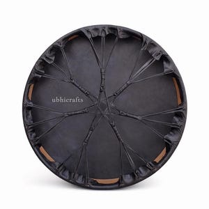 Puede incluir: Un tambor redondo negro con una superficie de piel estirada y un patrón tejido de cuerdas negras. El borde del tambor es visible, con un borde marrón claro. El texto "ubhicrafts" está impreso en la superficie del tambor.