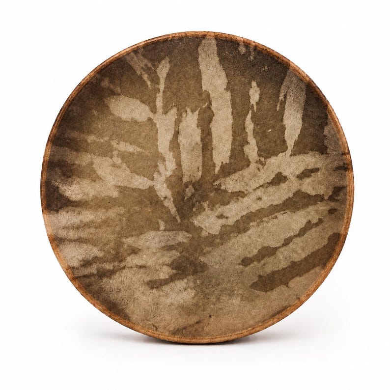 May include: A circular frame drum with a wooden rim and a natural skin head. The drum head features a mottled, earthy brown and beige pattern with organic, leaf-like shapes across the surface, creating a textured appearance on the drum skin.