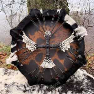 Può includere: Un tamburo rotondo, fatto a mano, con una faccia in pelle naturale con un motivo maculato marrone e nero. La cornice del tamburo è adornata da finiture in pelliccia bianca e nera. Un disegno intrecciato bianco e nero decora il centro.