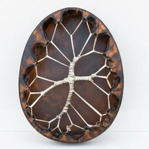 May include: An oval drum with a dark brown leather face and a lighter brown, stitched leather border. A white, tree-like design is woven across the drum's face, creating a unique, artistic instrument.