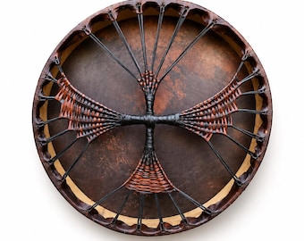 May include: A circular frame drum with a dark brown, mottled skin head. The back features a complex lacing system made of black cord and woven brown and black strips that form a cross-like pattern radiating from the center to the wooden rim.