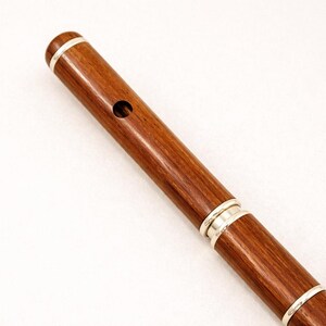 Puede incluir: Primer plano de una flauta de madera con detalles plateados. La flauta es de color marr&oacute;n oscuro con un acabado pulido. Bandas plateadas rodean los extremos y una secci&oacute;n de la flauta. Un solo agujero es visible cerca de la parte superior.