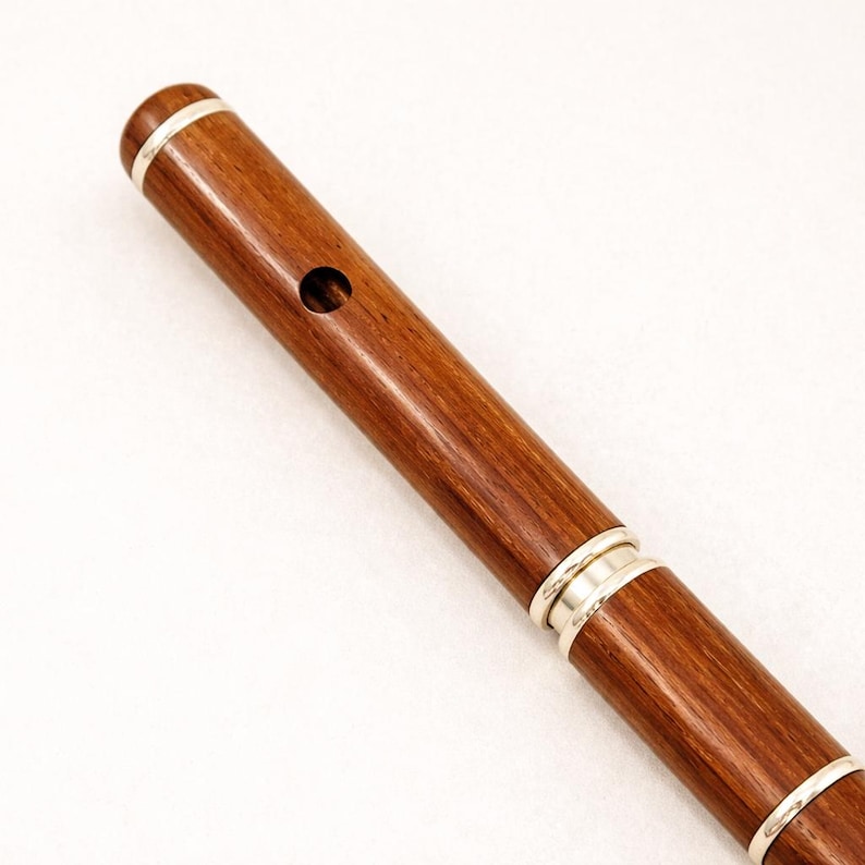 Puede incluir: Primer plano de una flauta de madera con detalles plateados. La flauta es de color marr&oacute;n oscuro con un acabado pulido. Bandas plateadas rodean los extremos y una secci&oacute;n de la flauta. Un solo agujero es visible cerca de la parte superior.