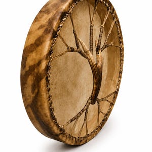 May include: A round frame drum with a light tan drumhead and a dark brown mottled wooden frame. The drum features a central handle shaped like a tree branch, connected to the frame by thin, light-coloured cord lacing around the entire perimeter.