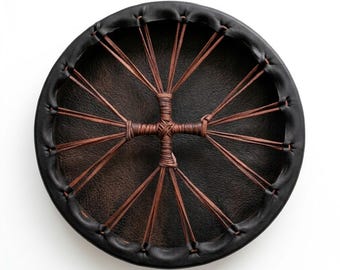 May include: A circular frame drum featuring a dark brown textured drumhead stretched across a black leather rim. The drumhead is secured by thin brown leather lacing that radiates from a central cross-shaped knot, creating a symmetrical pattern.