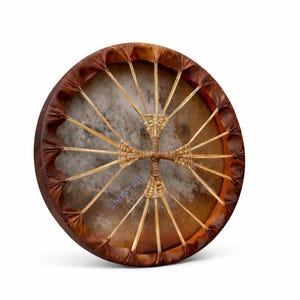 May include: A circular frame drum with a natural tan animal hide head. The drum features a dark brown leather rim and a complex lacing system made of thin, light-coloured cords that converge in the centre to form a woven cross-like pattern.