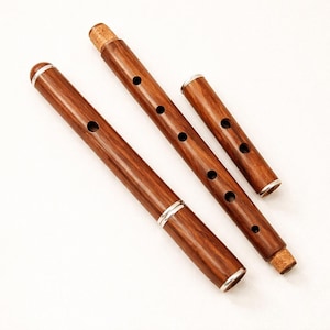 Puede incluir: Tres flautas de madera con un acabado marr&oacute;n oscuro y detalles plateados. Cada flauta tiene varios agujeros para los dedos y un tap&oacute;n de corcho. Las flautas est&aacute;n dispuestas en diagonal sobre un fondo blanco.