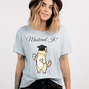 Op de afbeelding: Lichtblauw T-shirt met de tekst "Mastered It!" boven een cartoonkat die een afstudeerhoed draagt en een koffiekopje vasthoudt. De kat is bruin met bruine strepen en roze voeten.
