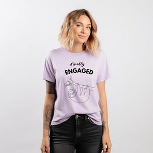 Peut inclure: T-shirt lilas avec l'inscription "Finally ENGAGED" en noir. Un dessin au trait d'un paresseux suspendu à une branche se trouve en dessous. Le t-shirt est de style col rond.
