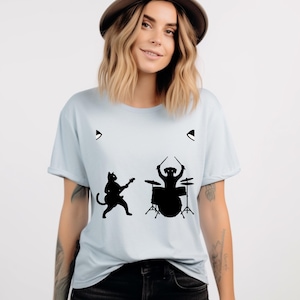 Peut inclure: T-shirt bleu clair avec une silhouette noire d'un chat jouant de la guitare et d'un chien jouant de la batterie. Le t-shirt a un col rond et des manches courtes. Le motif est centré sur le devant.