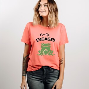 Könnte beinhalten: Korallfarbenes T-Shirt mit dem Text "Finally Engaged" über einer grünen Froschgrafik, die einen Ring hält. Das Shirt ist ein kurzärmeliger Rundhalsausschnitt.