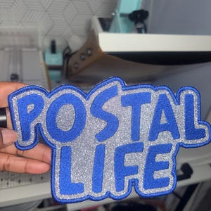 May include: A blue and silver patch with the words "POSTAL LIFE" in a stylized font. The patch has a blue border and a silver glittery center, creating a bold and eye-catching design. The patch is likely for clothing or accessories.