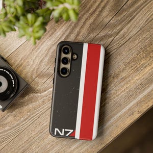 Puede incluir: Una funda de smartphone negra con un patrón de estrellas moteadas, una franja vertical blanca y una franja roja ancha. En la esquina inferior izquierda aparece el texto N7 en blanco. La funda tiene un recorte para triple cámara trasera.