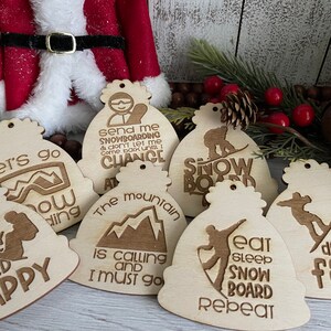 May include: Six wooden ornaments shaped like ski hats with various winter sports-themed sayings. The sayings include "Let's go snow boarding", "Don't worry board happy", "Send me snowboarding & don't let me come back until I chance at the mountain is calling and I must go", "Snow board", "Eat sleep snowboard repeat", and "My life is going downhill fast!"