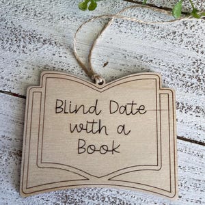 May include: A wooden ornament shaped like an open book with the text "Blind Date with a Book" engraved on it. The ornament is attached to a twine string.