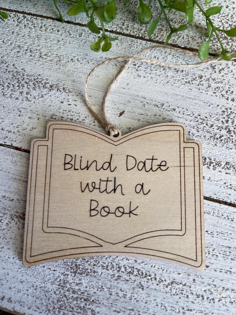 May include: A wooden ornament shaped like an open book with the text "Blind Date with a Book" engraved on it. The ornament is attached to a twine string.