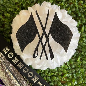 May include: A white flower with black glitter flags crossed in the center. The text "HOMECOMING" is on a black ribbon.