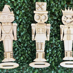 May include: Three wooden decorative figures in the shape of nutcrackers. One is a cat, one a skull, and the third a jack-o'-lantern. Each figure is light brown and stands on a small oval base, set against a green background.