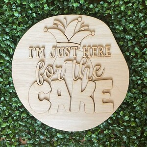 May include: A round wooden sign with the text "I'm just here for the cake" in a whimsical font. A jester's hat is above the text.