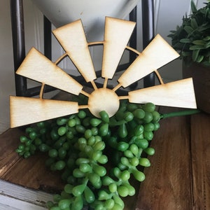 May include: A wooden cutout of a windmill with eight blades. The windmill is painted in a natural wood color and is sitting on a wooden surface.