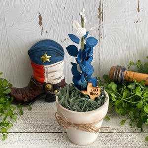 May include: A small white ceramic pot with a blue and white floral arrangement and a wooden Texas-shaped sign that reads "God Bless." A decorative cowboy boot and a wooden rolling pin are also visible.