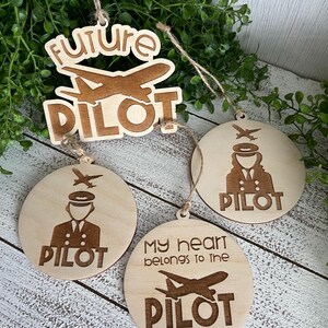 May include: Wooden ornaments with pilot-themed designs. One ornament reads "future pilot" with an airplane. Two round ornaments feature pilot figures and the word "PILOT". One round ornament says "My heart belongs to the PILOT".